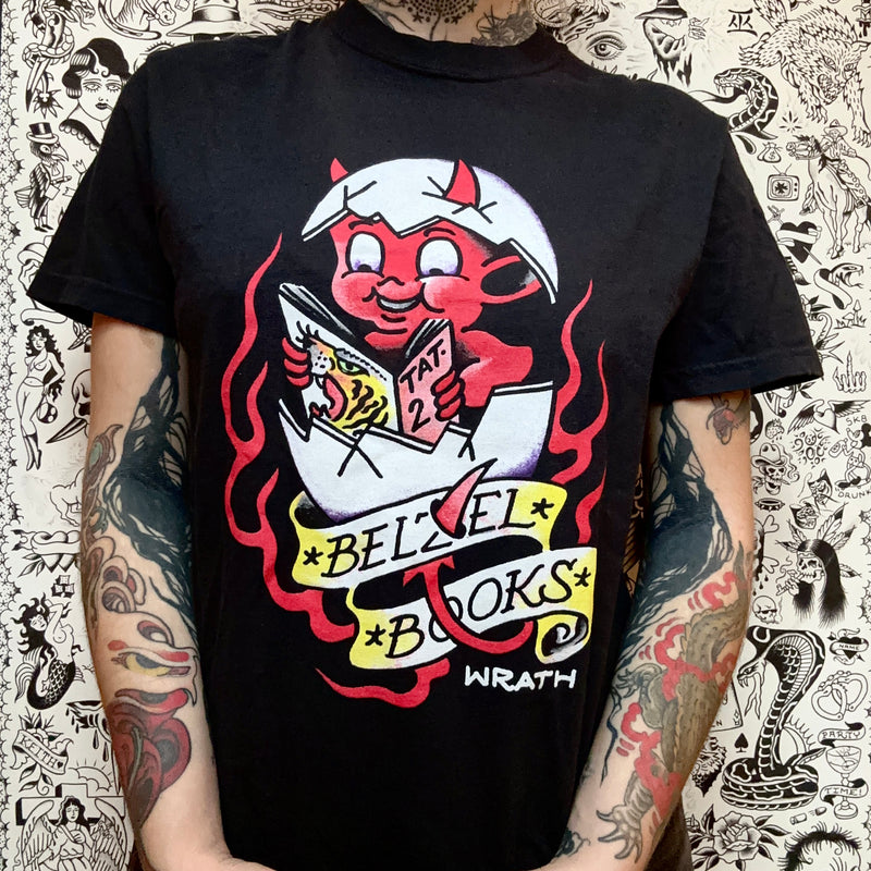 Front of a black t-shirt with a hot stuff devil inside of an egg, reading a magazine, with the belzel books banner at the bottom, screen printed on the front, and Wrath's signature at the bottom