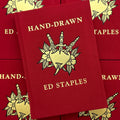 Front cover of Hand-Drawn by Ed Staples featuring a red book with gold text and design showcasing a heart with three daggers adorned by flowers.