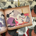 Inside pages of, 'Shunga' featuring a geisha woman in a purple robe laying down with a man over her about to insert his penis while he is touching her.