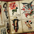 Inside pages of Ben Corday's Tattoo Travel Book featuring traditional designs in color showcasing an eagle with a 'death or glory' banner, a hand with playing cards, a rose, a Western-style woman, a skull with sailor uniform, and a pirate.