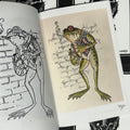 Inside pages of Subculture Frogs featuring a color illustration of a punk frog wearing a black vest, smoking a cigarette, and holding a covered bottle.