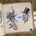 Inside page of Front cover of Jonathan Shaw's - Vintage Tattoo Flash Collection: Skulls Vol.1 showcasing two skulls, one skull is entwined with a snake and the other is wearing a black top hat and something sticking out of his mouth. 
